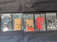 Five pairs of Halloween-themed earrings each sealed in individual plastic bags with black or red cards backing the earrings. Designs feature pumpkins, candy corn beads, cobwebs, witch hats, and bloody hatchets.