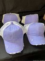 Four brand new lilac-colored baseball hats with distressed details, white mesh backs, and adjustable straps shown from front.