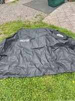 Full view of the black barbecue cover laid out on grass, showing its large size and brand text on the surface.