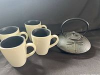 Image showing the full set of one cast iron teapot with dragonfly design and four ceramic mugs with cream exterior and black interior.