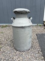 Full front view of full size gray metal milk can on gravel surface showing cylindrical body, side handles and fitted lid.