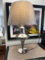 Full view of the lamp showing the grey fabric shade and reflective silver glass base on a round pedestal.