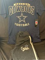 Photo of navy blue Dallas Cowboys T-shirt with large gold and white logo and text 'DALLAS COWBOYS FOOTBALL' and star, with tag attached.