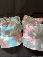 Front view of four ponytail hats showing two pink tie-dye and two blue tie-dye with distressed edges.