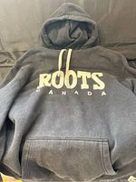 Front view of the black Roots sweatshirt hoodie showing the large white 'ROOTS CANADA' text on chest and hood with drawstrings.