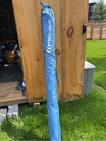 Blue Sport-Brella XL sun shelter folded inside a blue carrying bag leaning against a wooden wall outdoors.