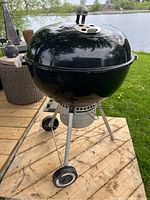 Front angled view of Weber charcoal grill showing lid with vent, steel legs and wheels near a lake backdrop.