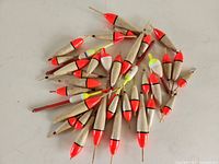 Multiple wooden fishing bobbers with painted tips laid out on a flat surface, predominantly red and some yellow tips, various sizes and shapes.