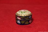 Front and side view of small round cloisonne ring box with floral needlepoint lid on red background.