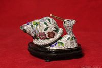Side view of enameled brass cloisonne snail figurine showing detailed floral enamel work on the snail shell and body, placed on a polished dark wooden base against a red background.