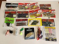 Photo of an assortment of fishing lures including soft plastic worms, paddle tails, tube worms, and small jig heads in packaging arranged on a table.