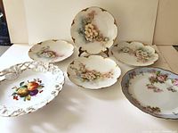 Seven porcelain plates arranged on a table: five Limoges plates with floral designs and gold trim, two Bavarian plates featuring fruit and floral patterns