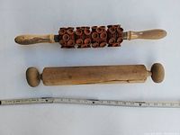 Photo showing two vintage wooden rolling pins on the floor next to a measuring tape for scale. One is a smooth wooden rolling pin with round handles, the other has a unique textured roller with rows of wooden knobs.
