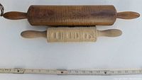Photo showing two wooden rolling pins, one large and plain, the other smaller with carved decorative patterns, along with a ruler for size reference.
