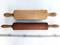 Photo of two vintage wooden rolling pins side by side, one light and one dark wood, with measuring tape below for scale.