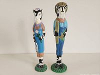 Front view of two standing wooden cow figurines, showing detailed painted clothing and facial features.