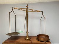 Wide view of antique brass balance scales with wooden base showing the two suspension dishes, brass on left and copper on right, with chain hooks and central brass column.