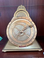 Front view of brass astrolabe clock showing circular plates with Roman numeral hour ring, zodiac engravings, month ring and central pointer with spherical counterweight on square base.