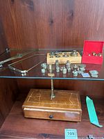 Photo showing wood box scale with scratches, various metal weights arranged on wooden base block, red plastic case with small weights, and a metal tweezer tool.