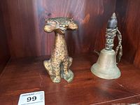 Wide angle photo showing both brass items, displaying the brass cat figurine with noticeable wear and the brass bell with decorative handle