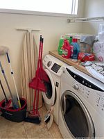 Photo showing various cleaning and laundry supplies including a striped ironing board, cleaning products on top of washer and dryer, brooms, mops, and dustpans clustered beside laundry machines.