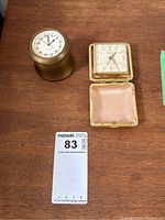 Two clocks on wooden table: brass Bulova desk clock with 24-hour dial and Selfix Timemaster travel alarm clock in leather case with cream square dial.
