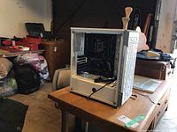 Partial assembled white computer tower on wooden table with plexiglass side panel, motherboard visible inside.