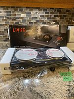 Living Basics double hot plate in original packaging with two burners visible, new and unopened condition, placed on a kitchen counter with tiled backsplash.