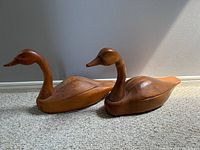 Two wooden duck carvings placed side by side on carpet causing visible shadows from sunlight. The ducks have carved contours and details.