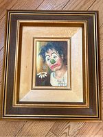 Framed oil painting showing full artwork of sad clown and flower with bee, with frame and wood floor background.