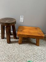 Photo of two small stools: one round dark wood stool with four legs and one square light pine stool with four legs, placed on carpet against a wall.