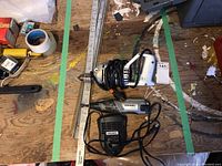 Photo displaying an electric drill with cord, a Dremel 8200 rotary tool with cord, charging adapter, and a folding measuring stick on wooden floor with green tape markings.