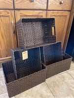 Photo showing five brown wicker storage bins stacked and arranged, three smaller bins inside one large bin on floor against kitchen cabinets.