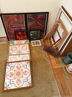 Photo showing framed needlepoint artworks and framed floral prints arranged on floor next to wooden picture frames and stained glass panel.