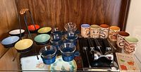 Full assortment of barware items including mugs, condiment bowls, bar tools, coffee cups with glass inserts, and drink book on wooden shelf.