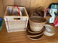 Collection of wicker baskets varying in shape and size including a large square basket and several round and oval baskets stacked together on a wooden surface.