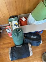Camping gear including Coleman air mattress packed in black/tan bag, green foam mattress roll, bellows foot pump box, fire extinguisher and orange sleeping bag.