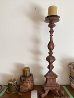 Tall wooden candle holder with a beige pillar candle placed on top, displayed against a wall on a wooden surface.