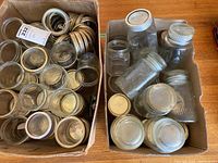 Two cardboard boxes containing assorted glass canning jars and metal lids/rings. One box contains more jars, the other mostly metal lids and some jars.