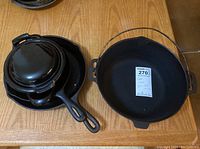 Four cast iron and enamel cookware pieces arranged on a wooden surface, including a large pot with metal handle, two fry pans, and an enamel pot with lid.