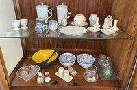 Photo showing upper shelf with variety of ceramic mugs with lids, miniature teacup, white plates, pottery pieces and some ceramic egg cups. Lower shelf with yellow enamel bowl with black utensils, two blue and white china bowls, small glass vases and heart-shaped glass dish.