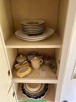 Cabinet shelf with stacked porcelain plates featuring floral and gold designs, beige teapots, pitchers and small pottery jars.