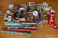 Photo showing an assortment of Christmas wrapping paper rolls, gift bags, glass ornaments, Christmas stockings and other holiday decor items spread out on a wooden table.
