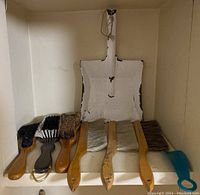 Photo showing metal dust pan with chipped white paint along edges, surrounded by seven brushes varying in size and handle type including wooden and plastic.