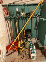 Photo showing various garden tools hanging on green wall and resting on floor: red snow shovel, weed eater, spades, pruning shears, yellow sprayer, bottles of Moss Out lawn chemical.