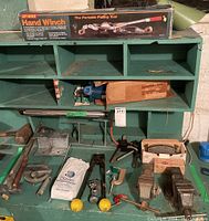 Wide view of hand winch box on top of green shelving unit with various clamps, pliers, and hardware below.