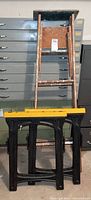 Photo showing the full wooden ladder standing upright with visible paint stains, leaning against metallic drawers, and the two black and yellow sawhorses positioned together.