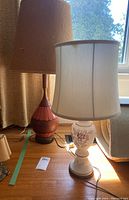 Two MCM style table lamps side by side on wooden floor near window, one white floral painted, the other reddish-brown with orange accents.