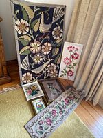 Six pieces of needlepoint art arranged on floor and leaning against wall, including framed wall hangings and a runner