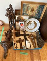 First image showing various religious statues, crucifixes, plates, framed pictures and cross stitch arranged in a shallow box on the floor.
